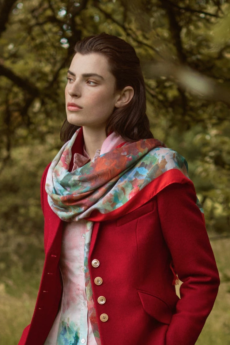 SCAPA AW25 Woman in a red coat and floral dress standing in a grassy field with trees.
